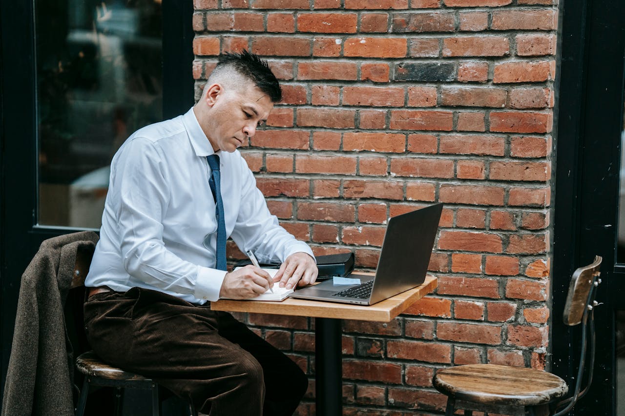 Man writing in a notebook beside a laptop at a small table, representing thoughtful essays and reflective writing